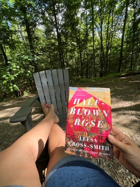 Picture of Pebbles' legs with feet on an adirondack chair. Holding the book Half Blown Rose by Leesa Cross-Smith