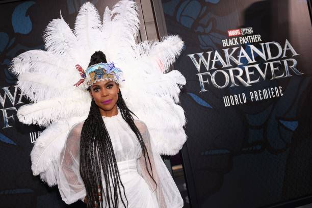 Hannah Beachler in a white outfit with white plumes surrounding her hear and shoulders in front of a Wakanda Forever background