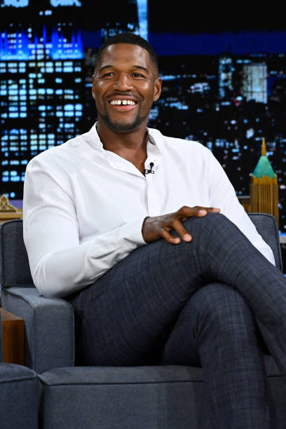 Michael Strahan in a white shirt crossing his legs on a talk show