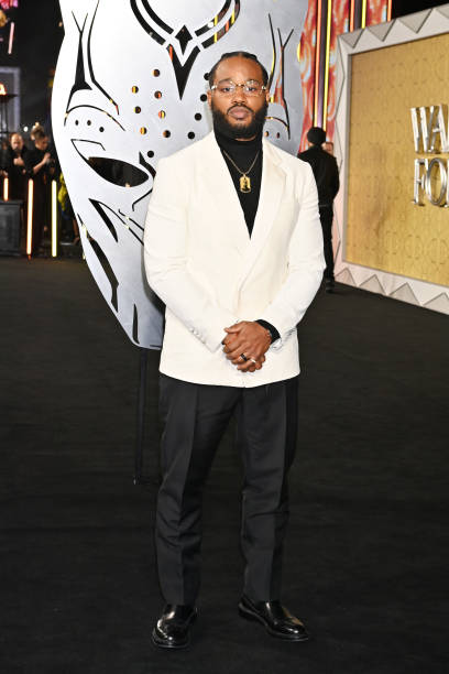 Ryan Coogler in a white jacket and black pants in front of a large black panther face mask