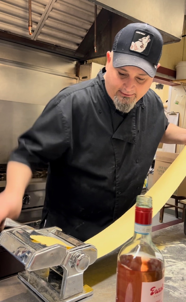 Picture of Chef Jason at Dave's Fresh Pasta rolling out dough in a pasta machine. He's wearing a black baseball hat and a black chef's jacket