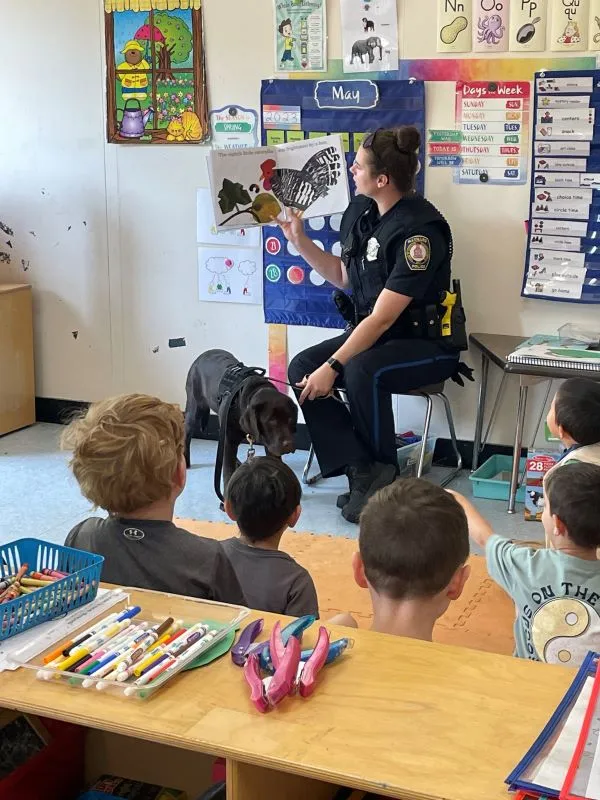 Dog handler Allison McCann holds up a book reading to a class of children, with chocolate lab Otis near her feet watching on.