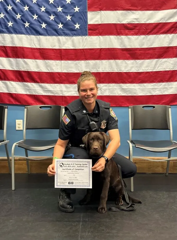 Picture of a chocolate lab comfort dog with his handler Allison McCann in uniform showing off the dogs training certificate. There is an American Flag in the background