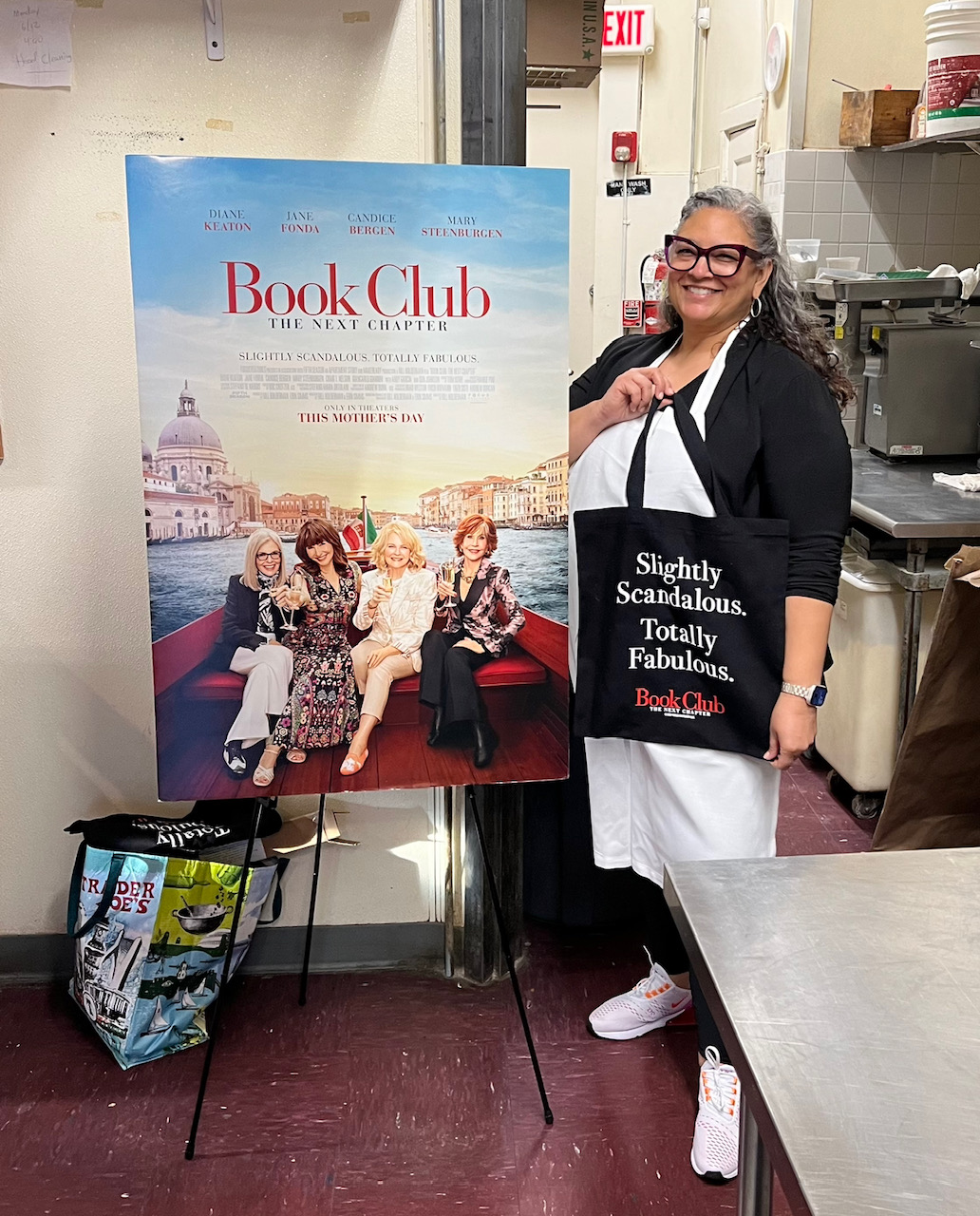 Pebbles in a restaurant kitchen next to a sign of the movie poster for Book Club - The Next Chapter. Also holding a bag that reads "Slightly scandalous. Totally Fabulous"