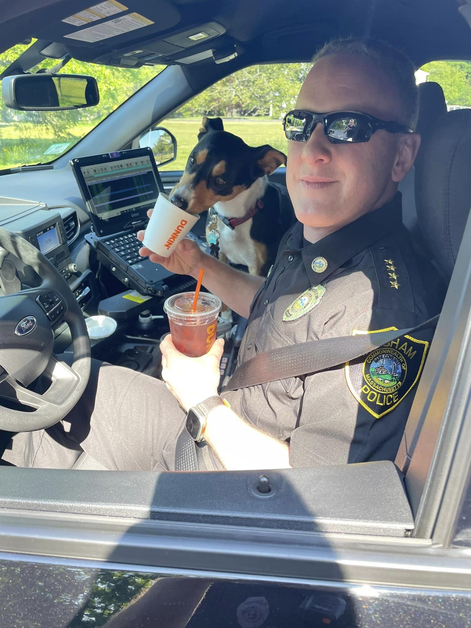 Police officer in the drivers seat of a car holding a iced drink from Dunkin. He also holds a smaller cup that a black, tan, and white dog is eating out of.