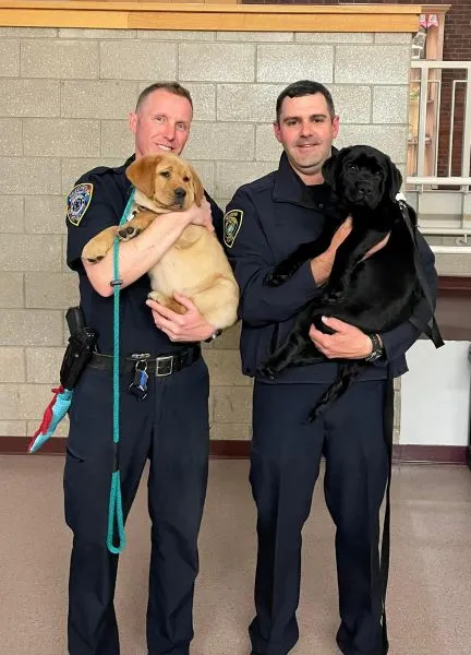 Two male police officers each hold one of the two comfort dogs of the department. One is a golden retriever and the other is a black lab