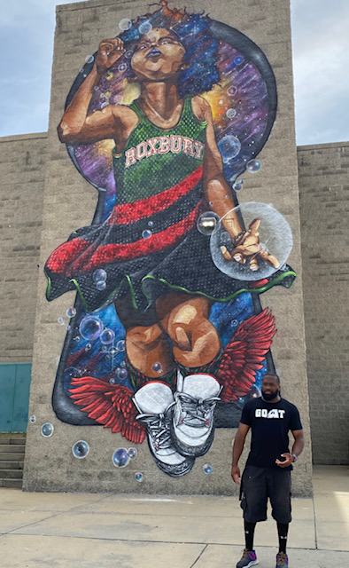 Artist ProBlak stands in front of his mural of a gilr blowing bubbles wearing a Roxbury jersey dress, and white high top sneakers with red wings.