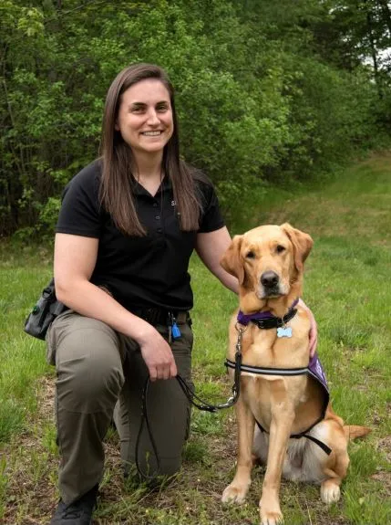 Female oficer with long brown hair kneeling with golden lab mix comfort dog Rico