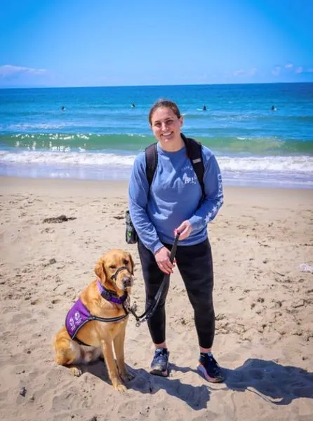 Female police officer wearing a blue sweatshirt and black leggings, with comfort dog Rico, a golden lab mix, enjoying a day at the beach.