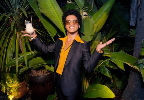 Picture of Bruno Mars in front of a large plant with a pina colada in his hand. Wearing a black suit, mustard-colored shirt and sunglasses