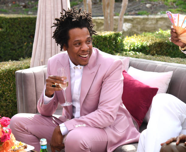 Picture of Jay-Z sitting, wearing a pink suit and holding a glass of champagne, smiling.