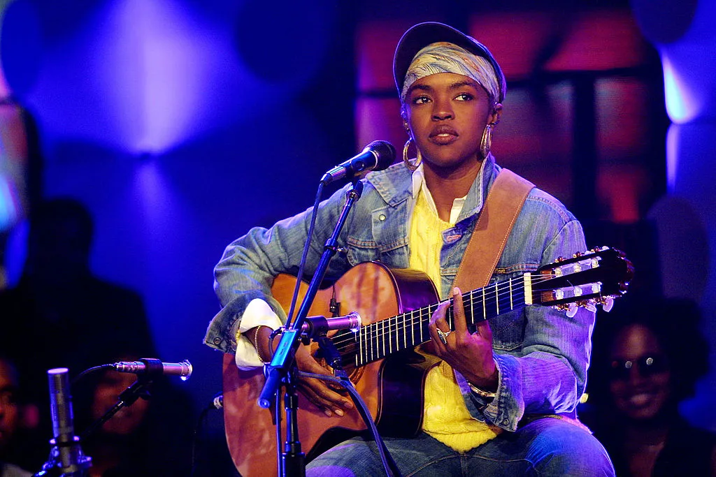 Lauryn Hill sitting on stage performing with her guitar and hair back in an all denim outfit
