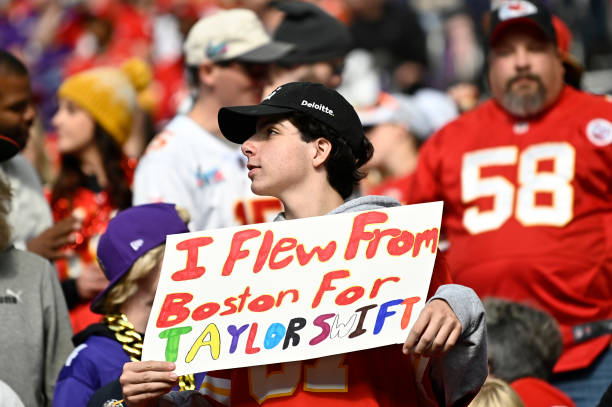 A guy wearing a black hat at a Chiefs football game holds a sign that says, "I flew from Boston for Taylor Swift.