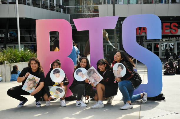 Five young woman in front of the letters BTS holding fans of their favorite group members faces