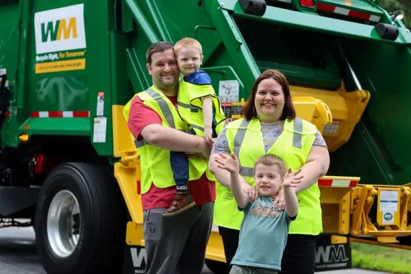 Father carries young son and mother stands behind other young son. All standing in front of a green and yellow garbage truck