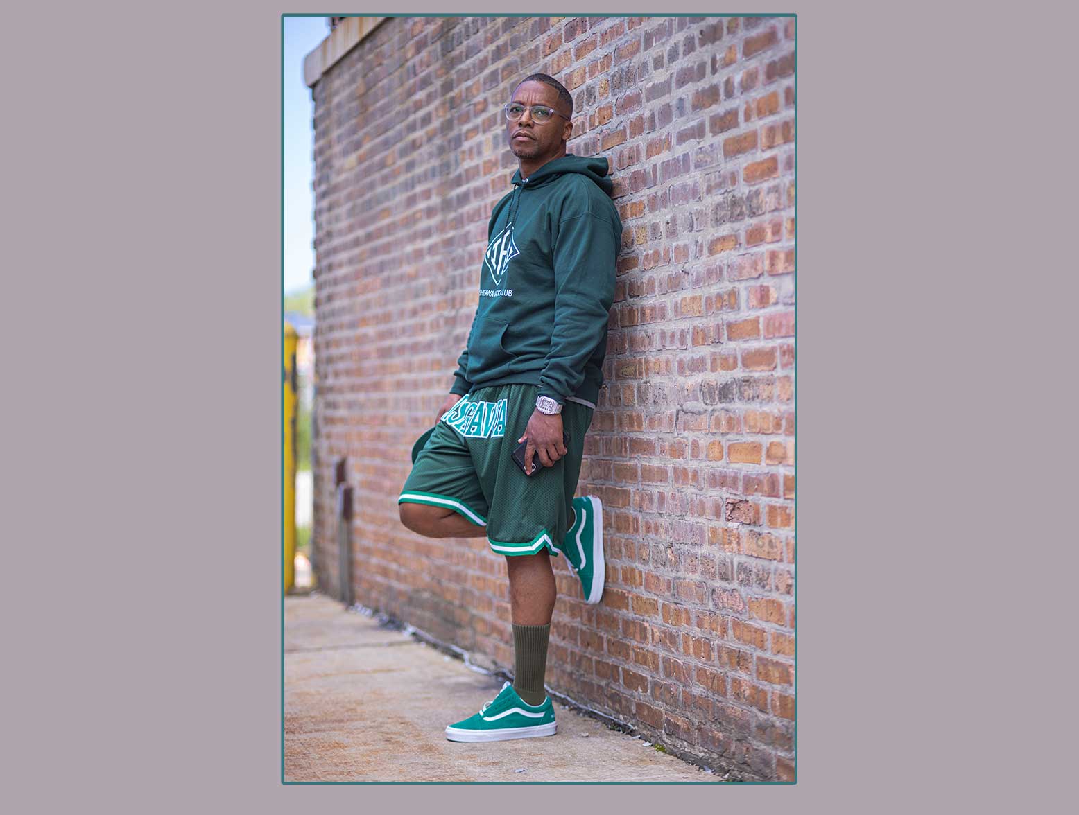 Lupe Fiasco leaning against brick wall in green sweatshirt and sneakers