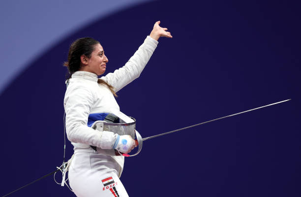 Side view of fencer Nada Hafez of Team Egypt holding her mask and foil in her right hand, and her left arm raised. 