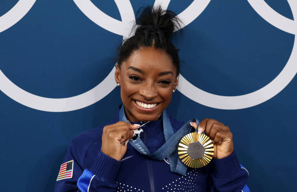 Close up of Simone Biles smiling, and holding up her gold medal and "goat" necklace. 