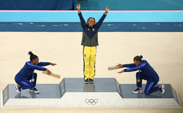 Simone Biles and Jordan Chiles bow down to Rebecca Andrade on the podium at the OIympics. 