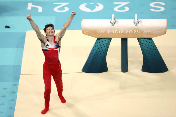 Olympic gymnast stephen nedoroscik in a red unitard uniform with his arms in the air standing next to a pommel horse.